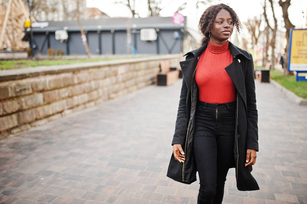 Portrait of a curly haired african woman wearing fashionable black coat and red turtleneck posing outdoor. - Foto, afbeelding