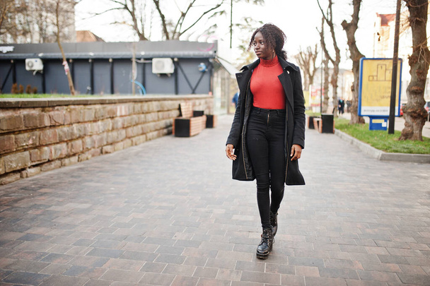 Portrait of a curly haired african woman wearing fashionable black coat and red turtleneck posing outdoor. - Φωτογραφία, εικόνα