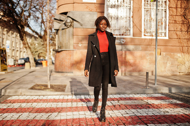 Portrait of a curly haired african woman wearing fashionable black coat and red turtleneck walking on zebra crossing. - Фото, изображение