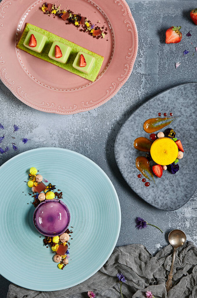 Tropical mousse desserts flat lay. Sweet candies and gourmet cakes served on luxury plates and handmade saucers top view. Confectionery desserts decorated with caramel smears, fresh fruits and petals - Zdjęcie, obraz