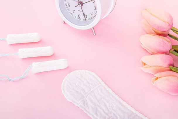 An alarm clock, sanitary pads, tampons and tulips on a pink background - Foto, afbeelding