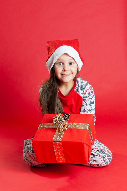 a happy girl in pajamas and a Santa Claus hat hands you a gift with a gold bow on a vivid background - Φωτογραφία, εικόνα