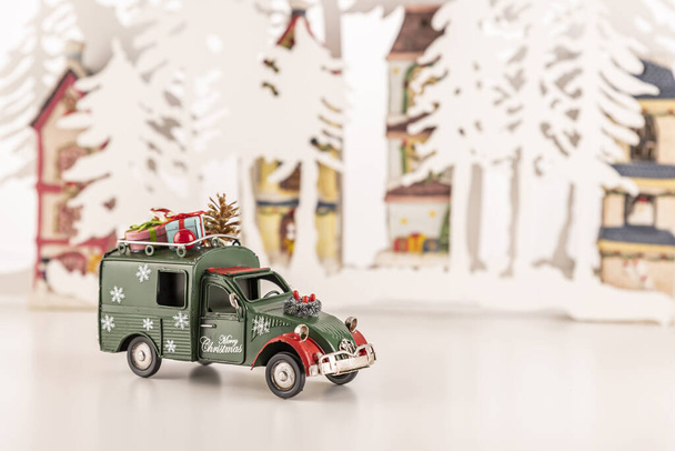 A small green toy car with ornaments on a white surface with trees and Christmas decorations in the background - Fotoğraf, Görsel