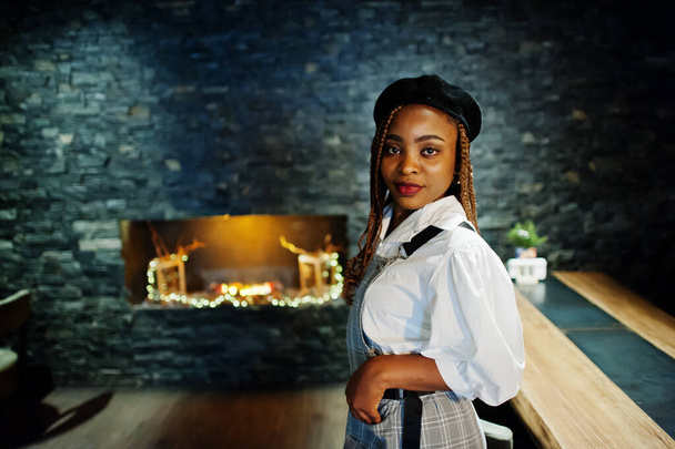 African american woman in overalls and beret in room with wooden table and fireplace. - Foto, afbeelding