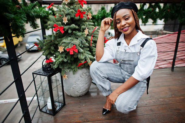 African american woman in overalls and beret posed in outdoor terrace with christmas decorations and new year tree. - Foto, Imagen