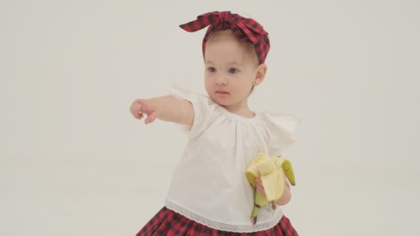 Kleines Mädchen mit kariertem Stirnband und Rock isst große Banane. Zeitlupe in Großaufnahme - Filmmaterial, Video