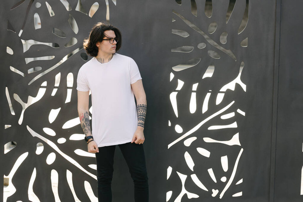 Hipster handsome male model with glasses wearing white blank t-shirt and black jeans with space for your logo or design in casual urban style - Φωτογραφία, εικόνα