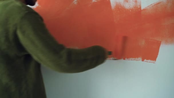 Un hombre se para cerca de la pared y pinta con un rodillo naranja. Copiar espacio. Concepto de renovación del hogar - Metraje, vídeo