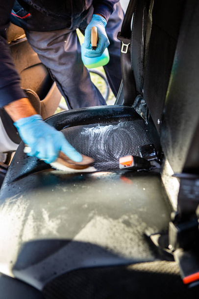 Wnętrze samochodu jest czyszczone. Pracownik używa specjalnego środka czyszczącego do czyszczenia fotelika samochodowego. Rozpyla chemikalia jedną ręką i rozmazuje pędzlem drugą.. - Zdjęcie, obraz