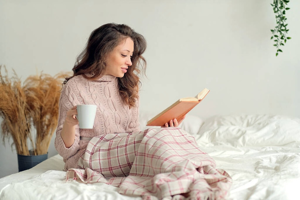 Jonge vrouw thuis zitten in de buurt van raam ontspannen in haar woonkamer lezen boek en het drinken van koffie of thee. - Foto, afbeelding