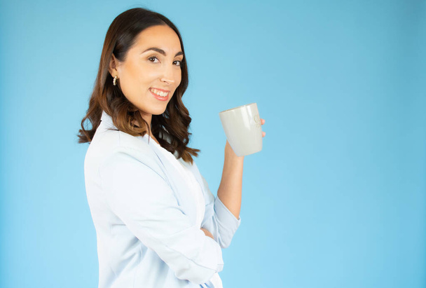 Jeune femme d'affaires buvant une tasse de café chaud sur fond bleu isolé avec une expression confiante sur le visage intelligent souriant - Photo, image