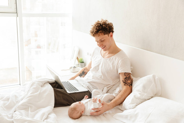 Happy young father playing with his little baby son while working on a laptop computer, laying in bed - Photo, image