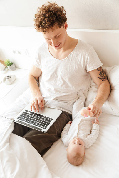 Happy young father playing with his little baby son while working on a laptop computer, laying in bed - Zdjęcie, obraz