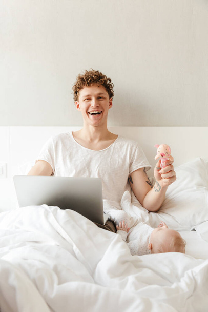 Szczęśliwy młody ojciec bawi się ze swoim małym synkiem podczas pracy na laptopie, leżąc w łóżku - Zdjęcie, obraz