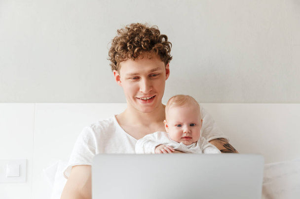 Happy young father playing with his little baby son while working on a laptop computer, laying in bed - Φωτογραφία, εικόνα