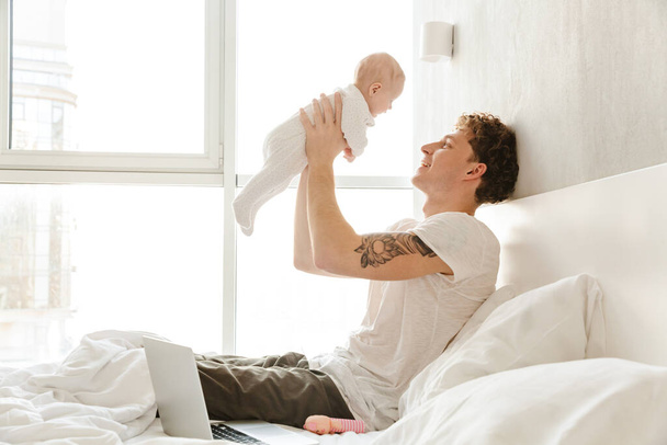 Happy young father playing with his little cutr baby son while laying in bed at home, embracing - Valokuva, kuva
