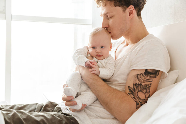 Happy young father playing with his little cutr baby son while laying in bed at home, embracing - Foto, Bild