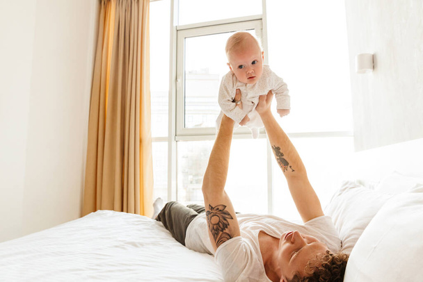 Happy young father playing with his little cutr baby son while laying in bed at home, embracing - Φωτογραφία, εικόνα