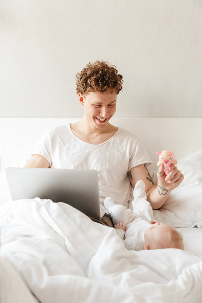 Onnellinen nuori isä leikkii pienen poikansa kanssa työskennellessään kannettavan tietokoneen kanssa, makaa sängyssä - Valokuva, kuva