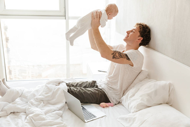 Happy young father playing with his little cutr baby son while laying in bed at home, embracing - Zdjęcie, obraz