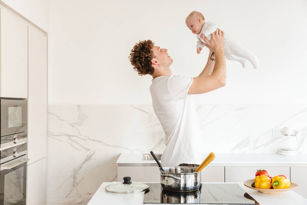 Smiling young attractive father cooking pasta while holding his baby son in the kitchen at home - Zdjęcie, obraz