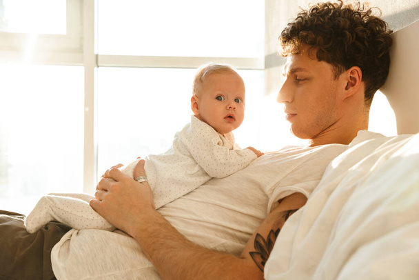 Happy young father playing with his little cutr baby son while laying in bed at home, embracing - Foto, Bild