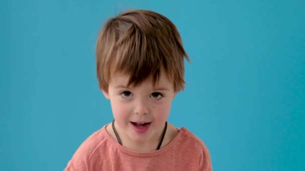 Niño con el pelo rubio asiente con la cabeza posando contra la pared azul - Metraje, vídeo