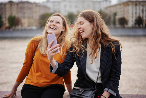 Дві красиві друзі молодої жінки гуляють разом по місту, використовуючи смартфон і радісно сміючись
 - Фото, зображення
