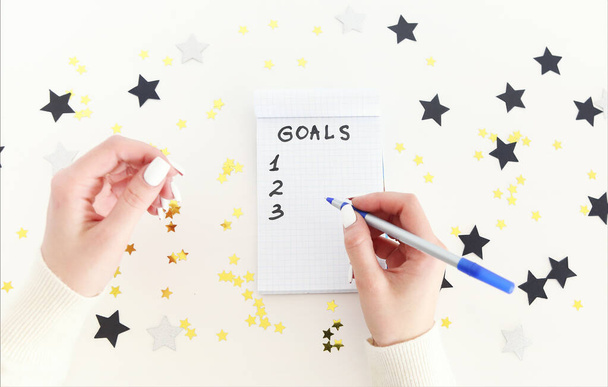 Main féminine avec stylo écriture objectifs du Nouvel An dans un carnet vide sur fond blanc avec confettis - Photo, image