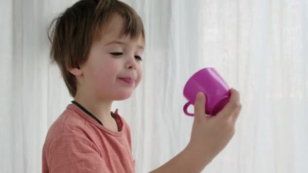 Uroczy mały chłopiec bawi się fioletowy kubek przez białą kurtynę - Materiał filmowy, wideo