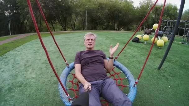 Man swinging on playground and talking at camera - Footage, Video