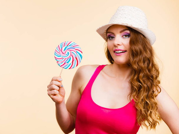 Beautiful woman wearing red tshirt summer hat holding big lollipop candy in hand. Sweet food fun concept. Studio shot on bright beige - Foto, imagen