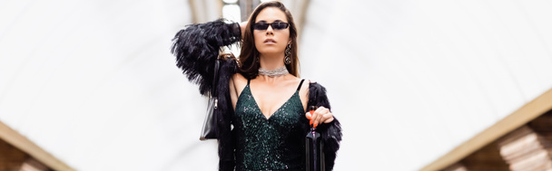 glamour woman in black dress, faux fur jacket and sunglasses posing with wine bottle at metro station, banner - Foto, Imagen