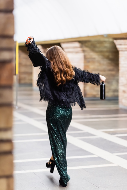 Uzun siyah lurex elbiseli zarif bir kadının arka planı metro istasyonunda el çantası ve şarap şişesiyle yürüyor. - Fotoğraf, Görsel