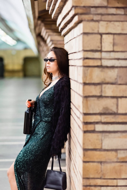ブラックルレックスのドレスとサングラスを着た魅力的な女性が地下駅でワインボトルと一緒に立っている - 写真・画像