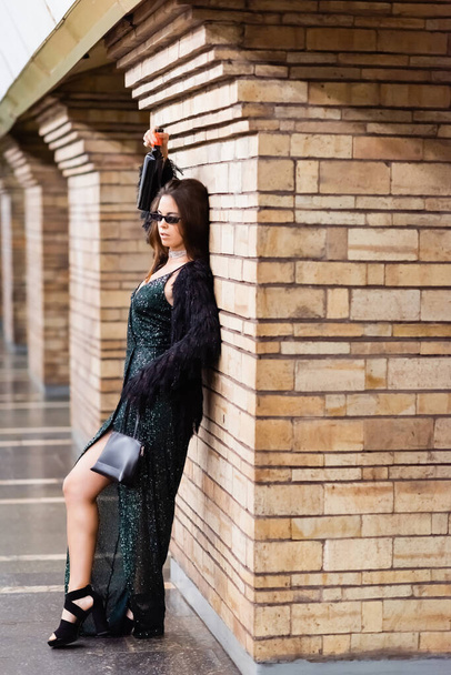 seductive woman in long black dress leaning on brick column while holding wine bottle in raised hand - Photo, Image