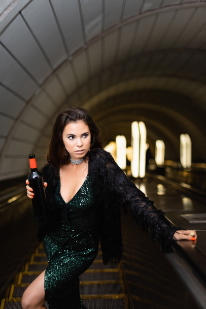 jovem mulher elegante em vestido lurex preto segurando garrafa de vinho enquanto olha para longe na escada rolante - Foto, Imagem