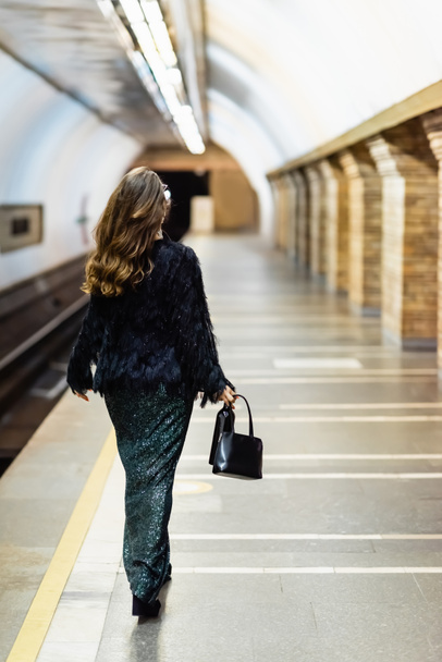 Rückansicht einer stilvollen Frau im langen Lurex-Kleid, die den U-Bahnsteig entlangläuft - Foto, Bild