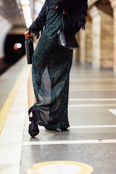 Uzun siyah elbiseli şık bir kadının metro platformunda elinde bir şişe şarapla görüntüsü. - Fotoğraf, Görsel