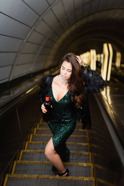 verführerische Frau im eleganten schwarzen Kleid, die das Haar berührt, während sie eine Weinflasche auf der Rolltreppe hält - Foto, Bild