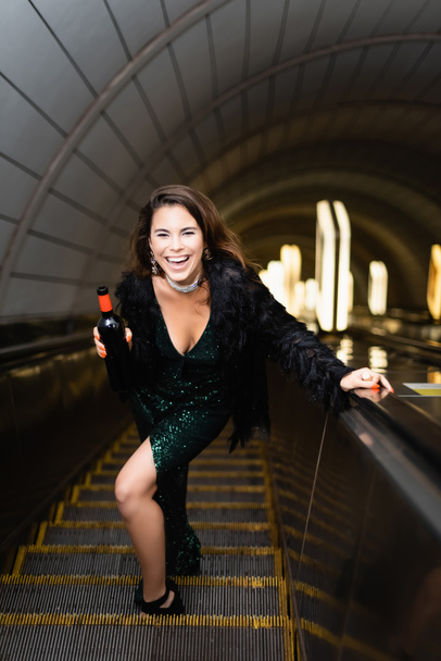 excited woman in elegant lurex dress laughing while holding wine bottle on escalator - Foto, Imagen