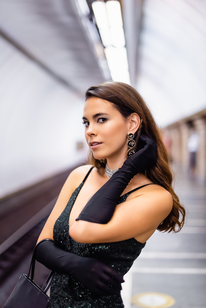 mulher sedutora em elegante vestido preto e luvas olhando para a câmera na plataforma do metrô - Foto, Imagem