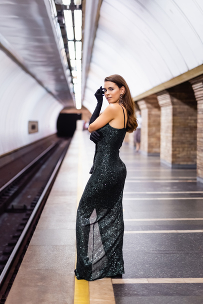 sensual woman in long black dress looking at camera while standing on underground platform - Photo, Image