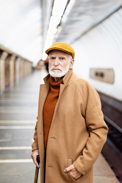 senior man in autumn cap and coat looking at camera while standing on underground platform - Фото, изображение