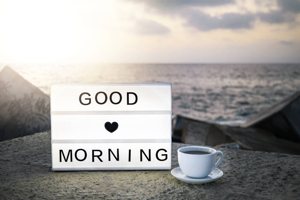 おはようございます。朝お茶やコーヒーのカップの横にあるライトボックスです。インスピレーションモチベーションライフ海と日の出を背景にライトアップされた箱にメッセージを引用 - 写真・画像