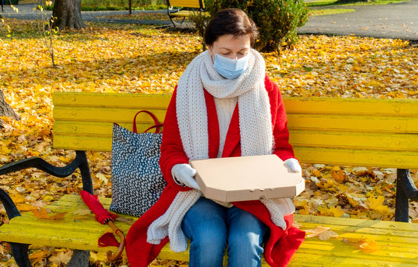 Женщина среднего возраста с защитной медицинской маской сидит на желтой скамейке в осеннем парке с коробкой для пиццы. - Фото, изображение