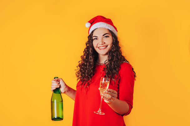 Young beautiful woman on yellow background, on her head she has santa hat. Woman holding bottle and glass in her hands. Christmas mood. Place for text - Photo, image