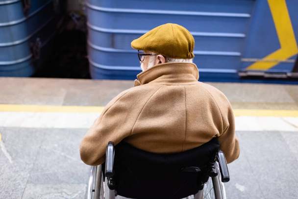 Rückansicht eines älteren behinderten Mannes im Rollstuhl, der Herbstkleidung trägt, in der Nähe eines verschwommenen Zuges auf einer Metrostation - Foto, Bild