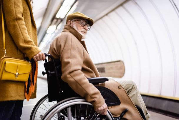 woman holding wheelchair of elderly handicapped man in autumn clothes on underground station - Foto, afbeelding