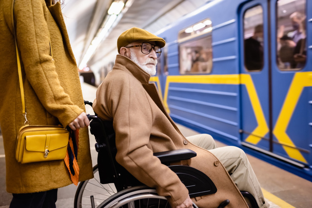 kobieta trzymająca wózek inwalidzki starszego niepełnosprawnego mężczyznę na podziemnej peronie w pobliżu rozmazanego pociągu - Zdjęcie, obraz